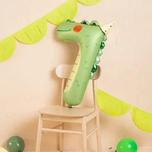 Peut inclure: Ballon en forme de dinosaure vert numéro sept avec un chapeau de fête. Le ballon est assis sur une chaise en bois avec une guirlande verte derrière.