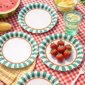 May include: A picnic scene with white paper plates featuring a green and white checkered border and orange trim. Fresh strawberries, watermelon, corn, potato chips, and a glass of lemonade are on a red and white checkered tablecloth.