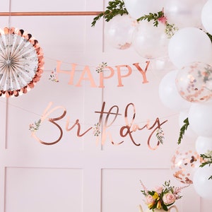 K&ouml;nnte beinhalten: Ros&eacute;goldene Happy Birthday Girlande mit floralen Akzenten. Die Girlande ist mit wei&szlig;em Garn aufgef&auml;delt und hat eine kursive Schriftart. Die Girlande h&auml;ngt an einer rosa Wand mit wei&szlig;en Ballons und Gr&uuml;npflanzen.