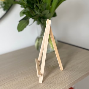 May include: A small wooden easel stand, made of natural light brown wood. The easel is standing on a light wood surface.