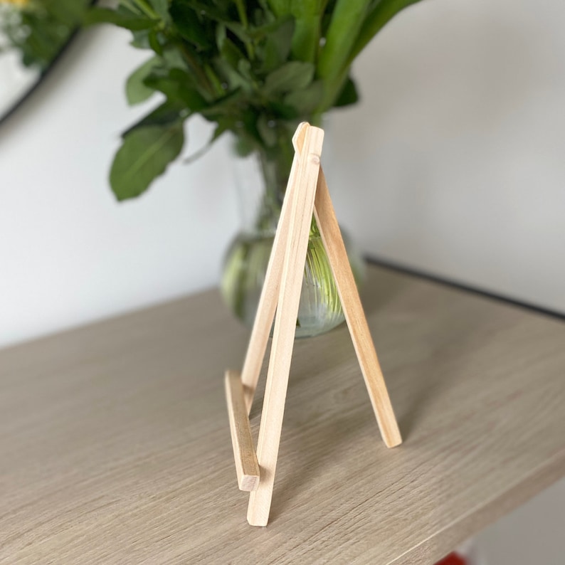 May include: A small wooden easel stand, made of natural light brown wood. The easel is standing on a light wood surface.