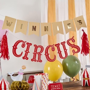 May include: A red and white circus themed party banner with the words "Winnie's Circus" in gold and red letters. The banner is strung between two red tassels and is decorated with gold and white striped paper.