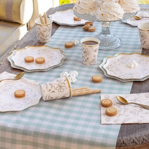 May include: A table setting with a light blue and white checkered tablecloth. The table is set with plates, cups, napkins, cookies, and meringue cookies. The plates and cups have a floral design with gold trim. A glass cake stand holds several meringue cookies.