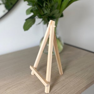 May include: A small wooden easel with a natural finish. The easel is made of four pieces of wood that are joined together to form a triangular shape. The easel is designed to hold a small painting or photograph.