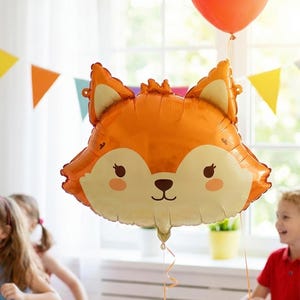 Puede incluir: Un gran globo de aluminio naranja con forma de cabeza de zorro, con una cara sonriente y detalles beige claro. Un segundo globo naranja flota encima. El fondo incluye niños y decoraciones de fiesta.