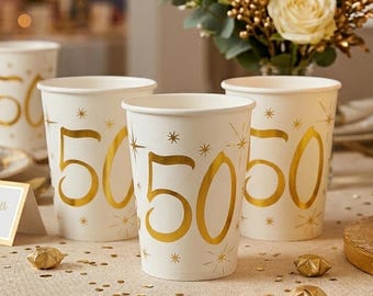 May include: Three white paper cups featuring a large metallic gold number 50 printed on the front, surrounded by small gold starburst designs. The cups are arranged in a group, showcasing the celebratory gold typography and star accents.