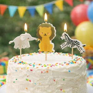 May include: A white iced cake decorated with colourful sprinkles, featuring three animal-shaped birthday candles: an elephant, a lion, and a zebra. The candles are lit with a flame.