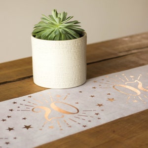 May include: White table runner with a gold foil design of the number 20 with stars and rays of light. The runner is on a wooden table with a small potted succulent plant.