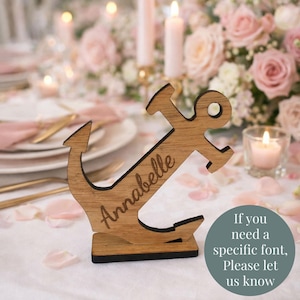 May include: Wooden anchor-shaped table place card holder with the name "Annabelle" engraved on it. The anchor is brown with a black base, set on a table with flowers and candles. Text on the image says "If you need a specific font, Please let us know."