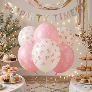 Può includere: Un gruppo di palloncini rosa e bianchi con un motivo a fiocchi ripetuto, posizionati davanti a uno striscione con la scritta PARTY TIME. I palloncini sono disposti sopra due alzatine per dolci con vari cupcake e macaron colorati.