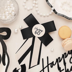 Pode incluir: Um laço listrado preto e branco com um círculo branco que diz "Happy 60". O laço está sobre um fundo branco com decorações pretas e brancas.