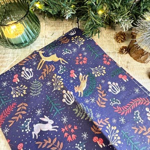Puede incluir: Papel de regalo azul oscuro con un diseño de bosque caprichoso. El patrón incluye ciervos dorados, un conejo blanco, setas y varias plantas en tonos verdes, rojos y marrones. Copos de nieve blancos están dispersos.