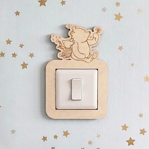 May include: A wooden light switch cover featuring a cartoon bear and a smaller character. The cover is a light brown color and is mounted over a white light switch. The background is a light blue wall with gold star stickers.