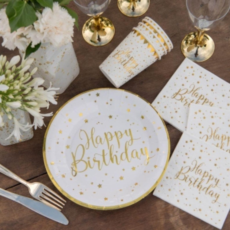 May include: White and gold birthday party supplies, including plates, napkins, and cups, are arranged on a wooden surface. The plate and napkins have "Happy Birthday" in gold script. Gold stars and a gold rim decorate the plate. Wine glasses and flowers are also present.