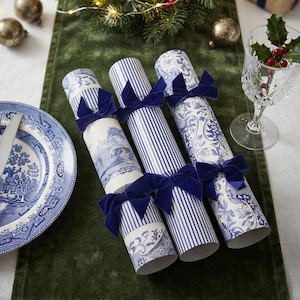 Op de afbeelding: Drie cilindervormige kerstcrackers met blauw-witte patronen. Twee crackers tonen gedetailleerde blauwe bloem- en landschapsontwerpen, terwijl de middelste verticale blauwe strepen heeft. Elke cracker is afgewerkt met donkerblauwe fluwelen strikken.