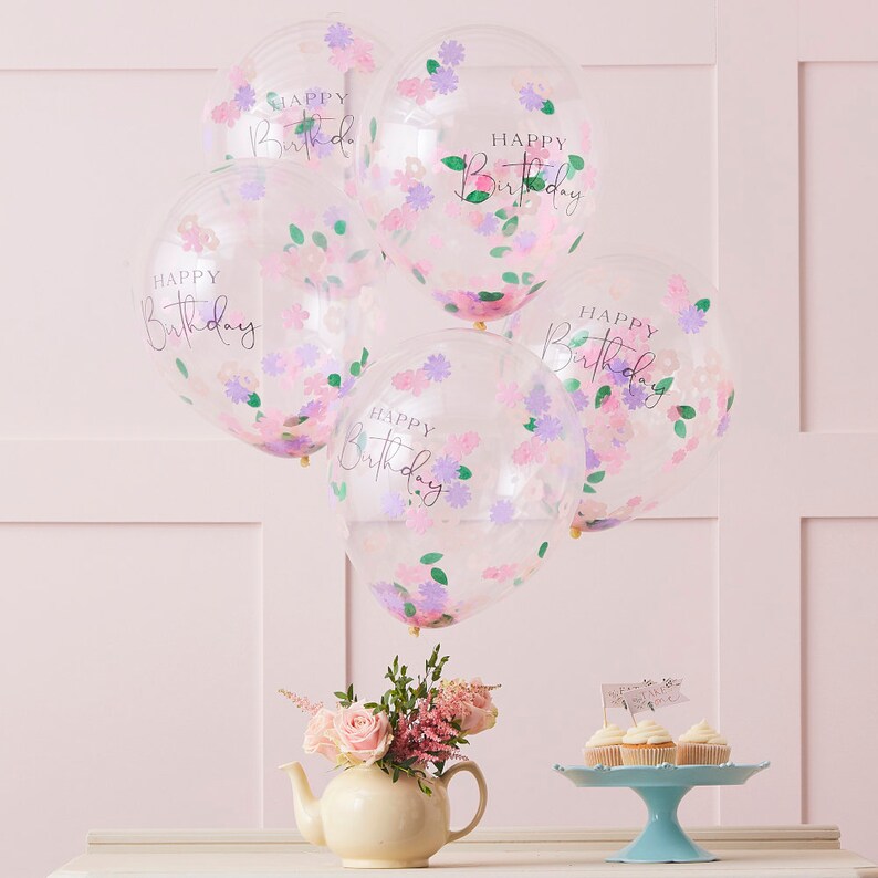 K&ouml;nnte beinhalten: F&uuml;nf transparente Ballons mit rosa und violettem Konfetti und dem aufgedruckten Text "Happy Birthday". Die Ballons sind zusammengebunden und schweben vor einer rosa Wand.