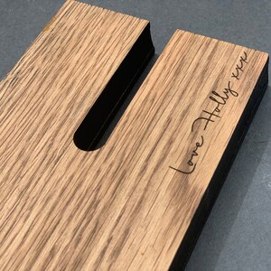 May include: Close-up of a wooden phone stand with a slot for the phone and the text "Love Holly xxx" engraved on the wood. The stand is made of light brown wood with a dark edge.