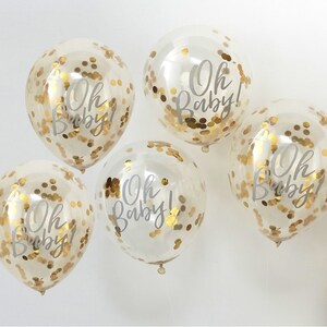 May include: Five clear balloons with gold confetti and the text "Oh Baby!" printed in white. The balloons are tied together with white string.
