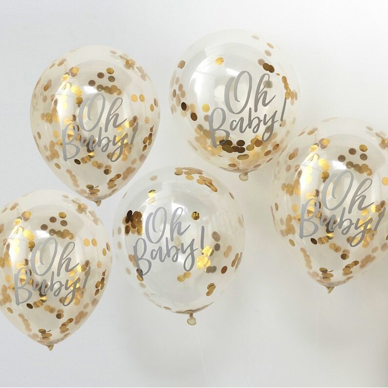 May include: Five clear balloons with gold confetti and the text "Oh Baby!" printed in white. The balloons are tied together with white string.