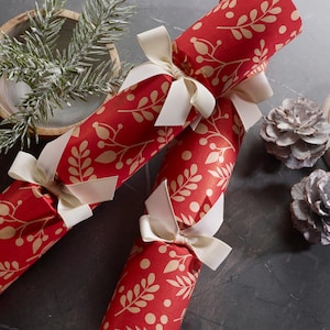 May include: Two cylindrical Christmas crackers wrapped in red paper with a repeating beige botanical leaf and berry pattern. Each cracker is cinched at both ends with a cream-colored grosgrain ribbon tied into a bow, securing the paper casing.