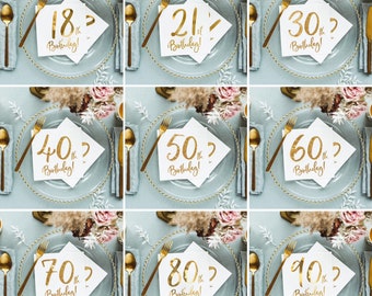May include: Nine gold-foil napkins with different numbers on them, from 18th to 90th, with the word "Birthday!" underneath each number. The napkins are placed on white plates with gold beaded edges, on a light blue background.
