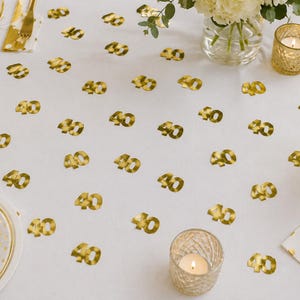 May include: A white tablecloth scattered with numerous small, metallic gold confetti pieces shaped like the number 40. The table setting includes a clear glass vase with white flowers, a gold-toned candle holder with a lit candle, and a gold fork.