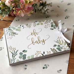 Può includere: Un libro degli ospiti bianco con un design floreale verde e oro e il testo "oh baby" in lettere dorate. Il libro ha un nastro bianco attaccato alla copertina.