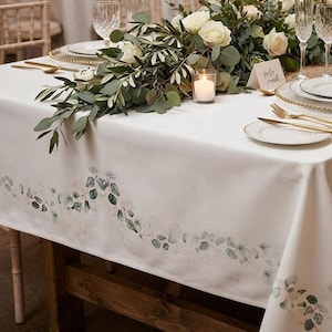 May include: A white rectangular tablecloth featuring a printed watercolour-style eucalyptus leaf garland pattern along the edges. The fabric is draped over a wooden table set with white plates, gold-toned cutlery, crystal glasses, and a candle.