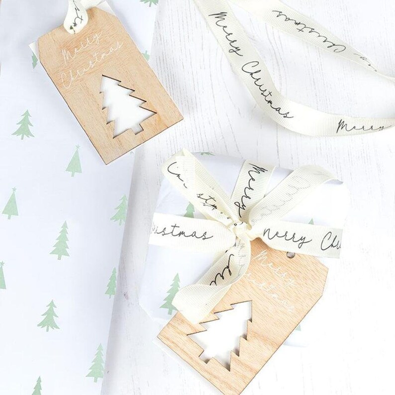 May include: Two wooden gift tags with a Christmas tree design and the text "Merry Christmas" on a white background with a white ribbon with the text "Merry Christmas" on it.