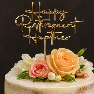 May include: A gold-colored cake topper that reads "Happy Retirement Heather" is placed on a cake decorated with peach and pink roses, white flowers, and green leaves. The cake has a light-colored frosting.