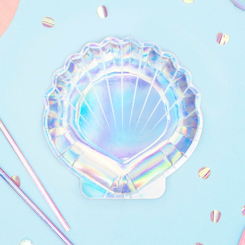 May include: Iridescent seashell-shaped paper plate with a scalloped edge. The plate reflects a rainbow of colors and has a fan-like design. Pink and silver straws and confetti are scattered around the plate on a light blue surface.