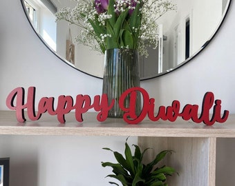 May include: Red wooden letters spelling "Happy Diwali" on a wooden shelf. A vase with white flowers and purple tulips is in the background.