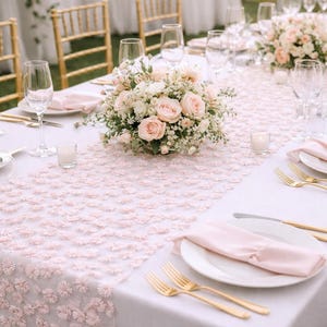 Peut inclure: Un chemin de table en dentelle florale rose clair avec un motif répétitif de petites fleurs en tissu en trois dimensions. Le chemin est posé sur une nappe blanche, accompagné d'assiettes blanches, de serviettes roses, de couverts dorés et de verres.