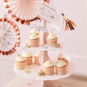 K&ouml;nnte beinhalten: Ein zweist&ouml;ckiger Cupcake-St&auml;nder mit einem Teekannen-Topper, auf dem "Let's Party Tea" steht. Der St&auml;nder ist mit rosa Punkten verziert und hat Cupcakes mit wei&szlig;em Frosting und F&auml;hnchen, auf denen "Take One" und "Yum Yum" steht.