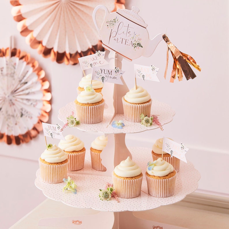 K&ouml;nnte beinhalten: Ein zweist&ouml;ckiger Cupcake-St&auml;nder mit einem Teekannen-Topper, auf dem "Let's Party Tea" steht. Der St&auml;nder ist mit rosa Punkten verziert und hat Cupcakes mit wei&szlig;em Frosting und F&auml;hnchen, auf denen "Take One" und "Yum Yum" steht.