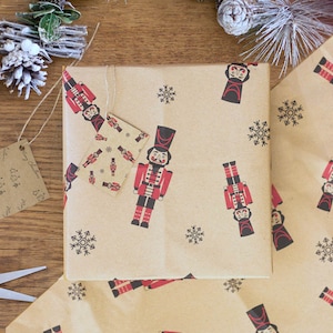 Op de afbeelding: Bruin inpakpapier met een herhalend patroon van notenkrakers en sneeuwvlokken. De notenkrakers dragen rode en zwarte uniformen met witte accenten. De sneeuwvlokken zijn zwart.