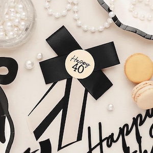 May include: A black ribbon with a circular sticker that reads "Happy 40" is the focal point of this image. The ribbon is set against a backdrop of pearls, macarons, and a black and white striped design, suggesting a celebratory theme.