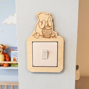 May include: Wooden light switch cover with a laser-cut design of Winnie the Pooh and a honey pot. The cover is rectangular with a switch cutout, mounted on a light blue wall. The word "HONNY" is visible on the honey pot. The design is suitable for a child's room.