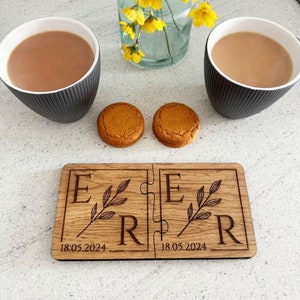 Puede incluir: Un juego de posavasos de madera con forma de rompecabezas con las letras "ER" y la fecha "18.05.2024" grabadas. El juego de posavasos está sobre una mesa con dos tazas de té y dos galletas de jengibre.