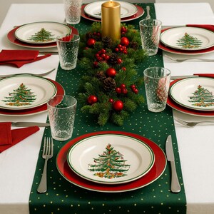 May include: A festive Christmas table setting with plates, glasses, and silverware. Each plate features a Christmas tree design. The table is decorated with a green runner, red napkins, and a centerpiece of greenery, red berries, and a gold candle.