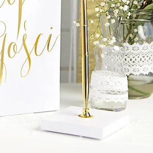 May include: A gold pen on a white square base. The pen is positioned in front of a white sign with gold script. In the background are glass vases with white lace and flowers, creating an elegant, decorative theme.
