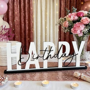 May include: A freestanding sign featuring the word HAPPY in large white block letters and the word Birthday in black cursive script, mounted on a black base. The sign is displayed on a table with a rose gold runner, candles, and flowers.