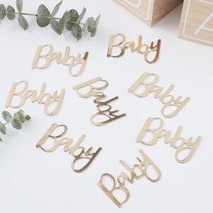 May include: Gold confetti with the word "Baby" written on each piece. The confetti is scattered on a white surface.