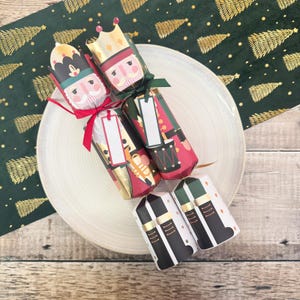 May include: A collection of Christmas crackers with nutcracker designs, arranged on a white plate. The crackers feature red, green, black, and gold accents, with blank tags for personalization. A green and gold Christmas tree runner is in the background.