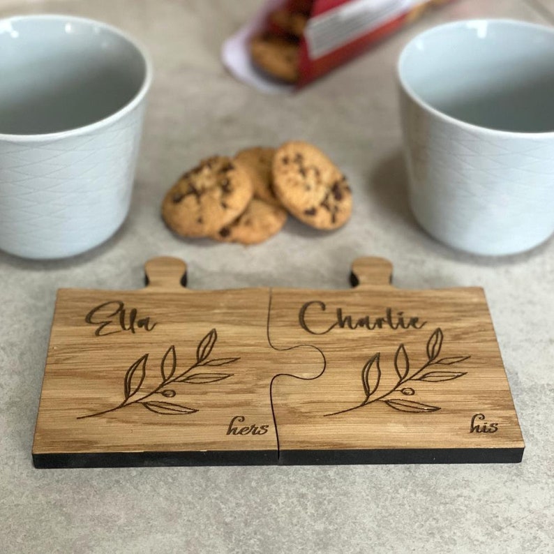 May include: Two wooden puzzle piece coasters with the names "Ella" and "Charlie" engraved on them. The coasters are joined together and have a black border. The words "hers" and "his" are also engraved on the coasters. The coasters are sitting on a table with two mugs and some cookies.