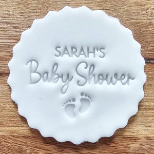 May include: A white, scalloped-edge disc with the text "SARAH'S Baby Shower" and two baby footprints. The text and footprints are embossed into the disc. The disc is on a wooden surface.
