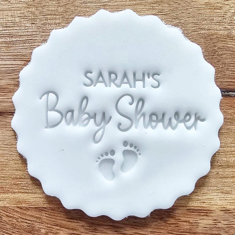 May include: A white, scalloped-edge disc with the text "SARAH'S Baby Shower" and two baby footprints. The text and footprints are embossed into the disc. The disc is on a wooden surface.