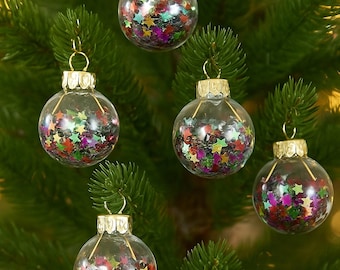 May include: Clear glass Christmas ornaments filled with colorful star-shaped confetti. Each ornament has a gold-colored cap and is hanging on a green Christmas tree branch. The confetti includes red, green, yellow, and purple stars.
