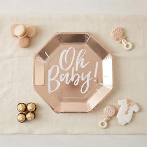 May include: An octagonal rose gold paper plate with the white text Oh Baby! printed in the center. The plate is surrounded by four round beige macarons, four gold foil-wrapped chocolates, two rattle-shaped cookies, and one onesie-shaped cookie.
