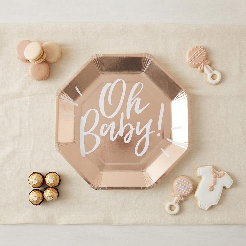 May include: An octagonal rose gold paper plate with the white text Oh Baby! printed in the center. The plate is surrounded by four round beige macarons, four gold foil-wrapped chocolates, two rattle-shaped cookies, and one onesie-shaped cookie.
